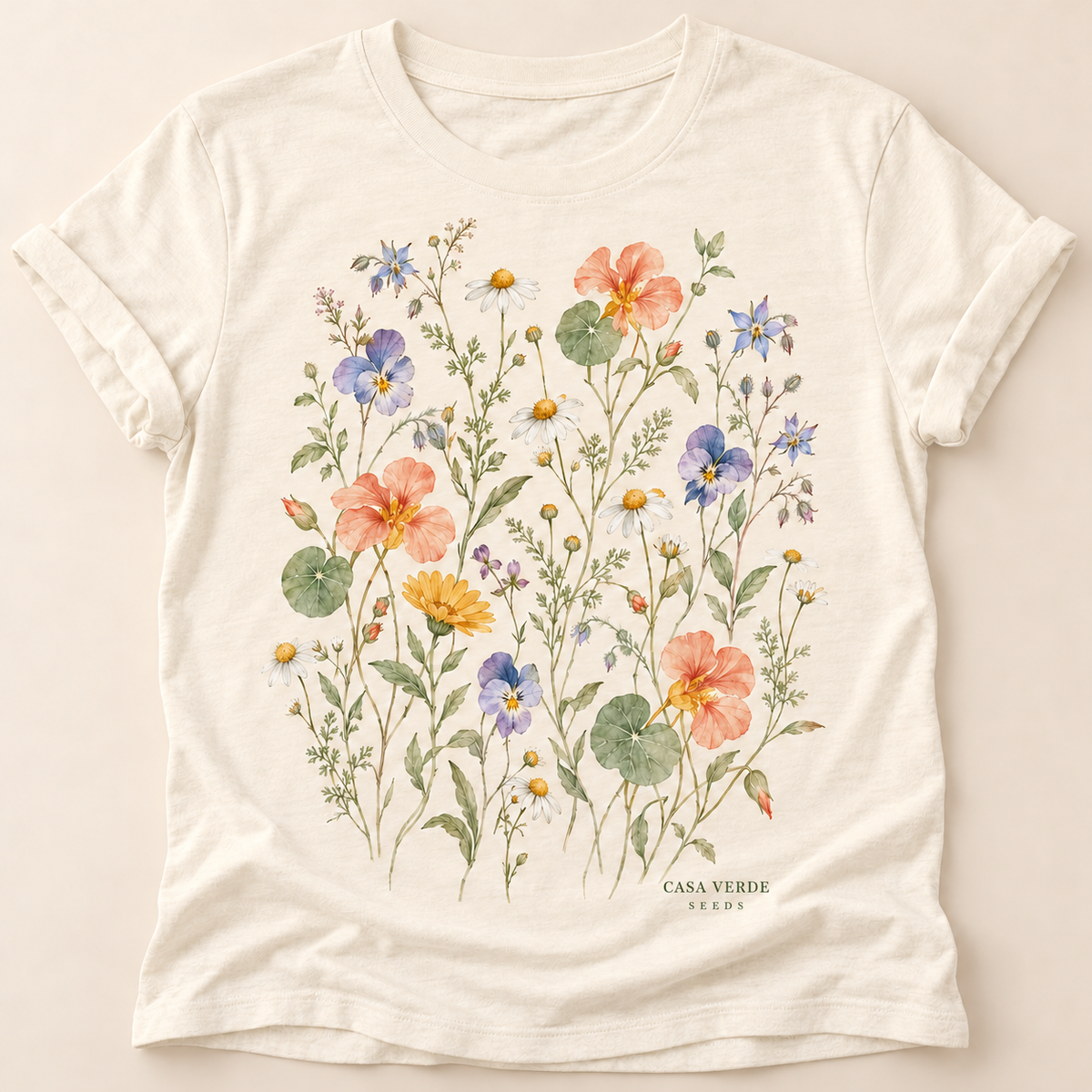 Ivory-toned T-shirt preview with a colourful watercolour edible-flower garden on the front With a small CASA VERDE SEEDS signature printed below the artwork. Design preview; final garment pending.