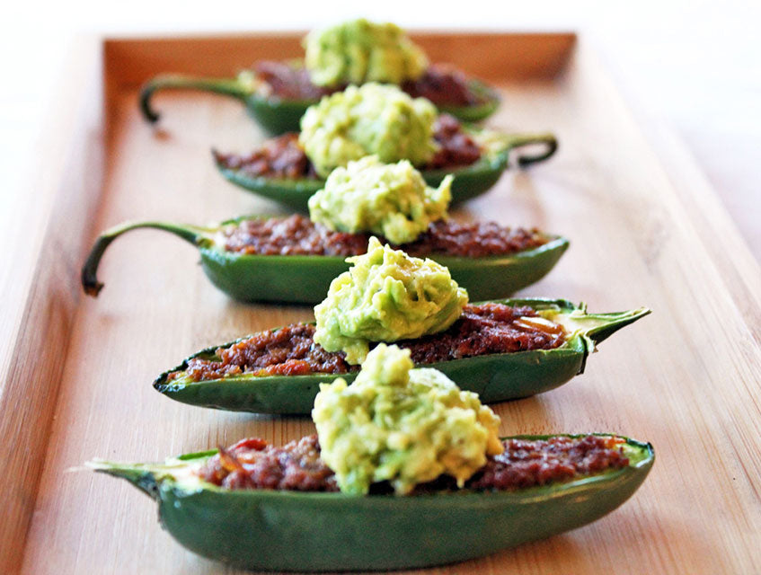nadapenoJalapeno halves filled with a red filling and topped with guacamole on a wooden board.