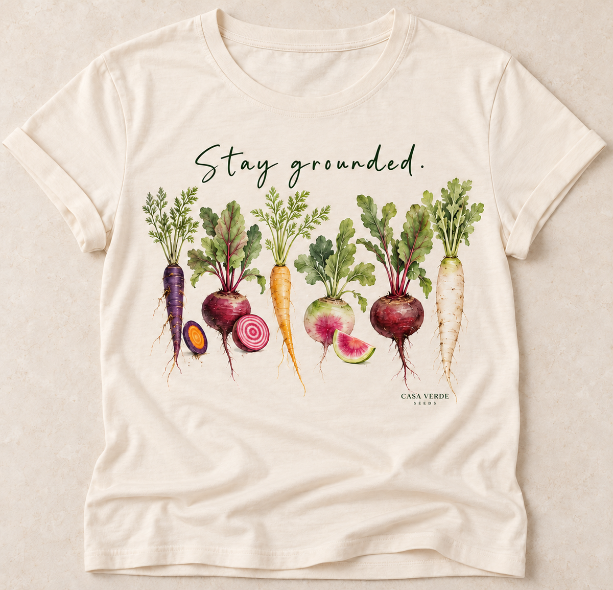Ivory-toned T-shirt preview with dark green handwritten Stay grounded., six watercolour heirloom roots and a small Casa Verde Seeds signature.